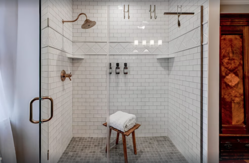 A modern shower with glass walls, white tile, a wooden stool, and three shower fixtures on the wall.