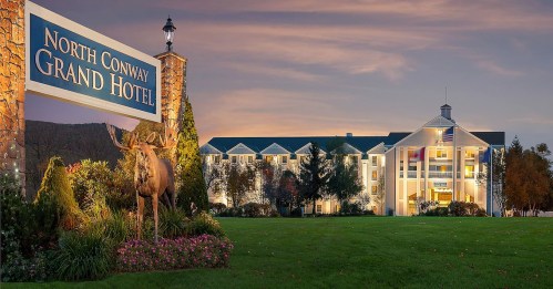 Sign for North Conway Grand Hotel with a stone structure and a moose statue in front, set against a sunset sky.