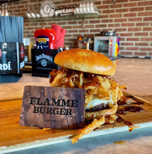 A delicious burger topped with crispy onions, served on a wooden platter with a Flamme Burger gift card.