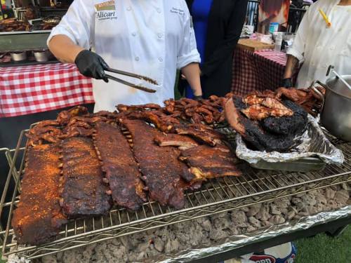 A chef uses tongs to serve a variety of smoked meats, including ribs and sausage, on a grill with a checkered tablecloth backdrop.