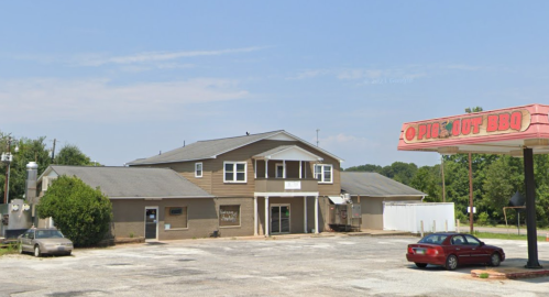 A brown building with a sign for "Opie's Out BBQ" in a parking lot, surrounded by trees and a clear blue sky.
