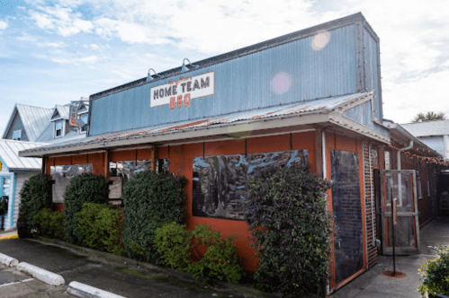 Exterior of Home Team BBQ restaurant with a metal roof and greenery, under a blue sky.