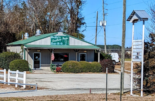 A small barbecue restaurant with a green roof and sign, surrounded by bushes and trees on a sunny day.