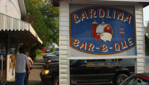 Sign for Carolina Bar-B-Que with a pig illustration, surrounded by parked cars and a person walking by.