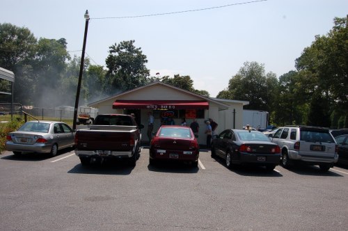 A barbecue restaurant with a red awning, surrounded by parked cars and people entering the building on a sunny day.