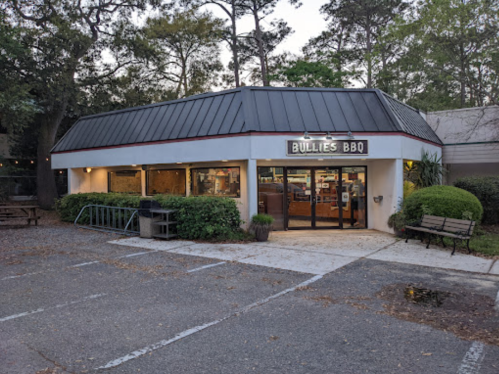 Exterior of Bullies BBQ restaurant, featuring a modern design, large windows, and outdoor seating in a wooded area.
