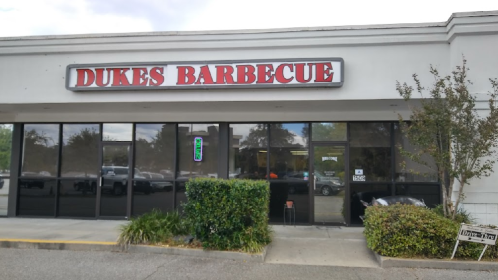 Exterior of Dukes Barbecue restaurant with a sign and large windows, surrounded by greenery and parking lot.