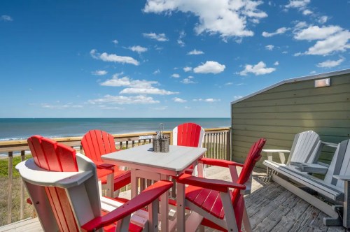 A seaside deck with red and white chairs, a table, and a view of the ocean under a blue sky with fluffy clouds.