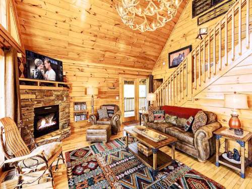 Cozy wooden cabin interior with a fireplace, comfortable seating, and a staircase, featuring rustic decor and warm lighting.