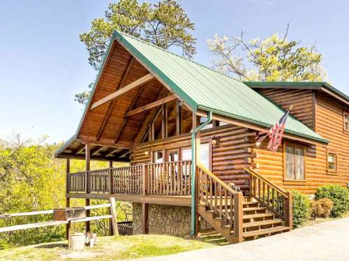 A rustic log cabin with a green metal roof, wooden porch, and American flags, surrounded by trees and blue sky.