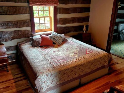 Cozy bedroom with a wooden log wall, a quilted bedspread, and a window letting in natural light.