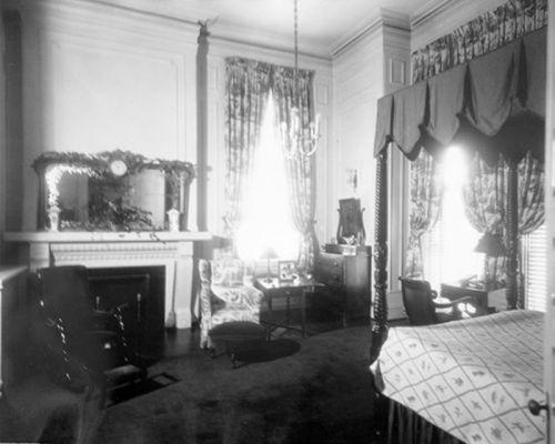 A vintage room with a fireplace, elegant curtains, a chandelier, and a table with a patterned cloth.
