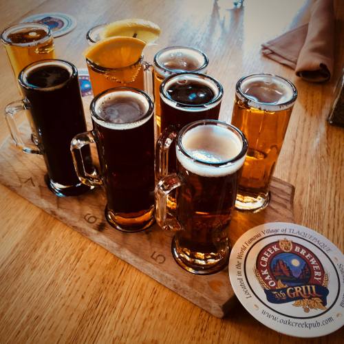 A wooden flight board with eight small beer glasses, featuring various colors and a lemon slice on one.