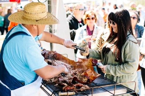 A vendor serves barbecue to a woman at a busy outdoor food festival, with a crowd in the background.