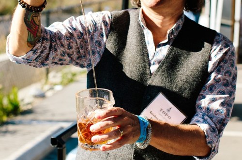 A person in a patterned shirt and vest stirs a drink in a glass, showcasing a colorful tattoo on their arm.