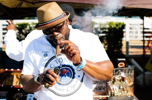 A man in a straw hat smokes a cigar, wearing a white shirt with a cigar logo, surrounded by a festive outdoor setting.