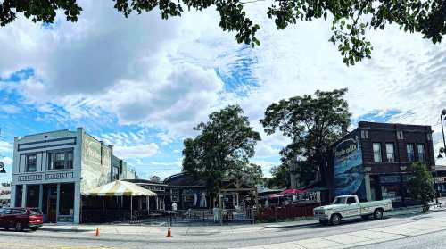 A street view of colorful buildings with outdoor seating, under a partly cloudy sky. A vintage truck is parked nearby.