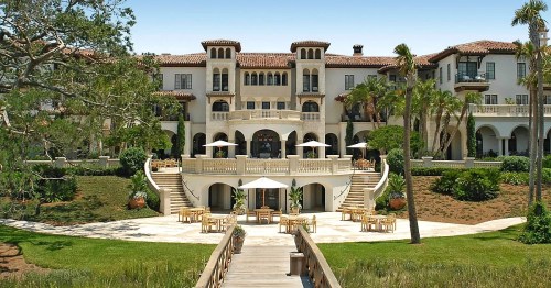 A grand Mediterranean-style mansion with a large terrace, surrounded by lush greenery and palm trees.