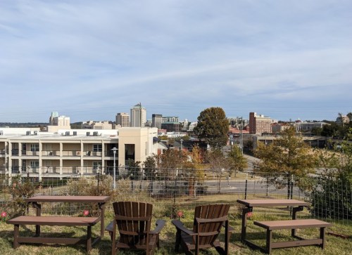 A view of a city skyline from a grassy area with wooden benches in the foreground and trees in the background.