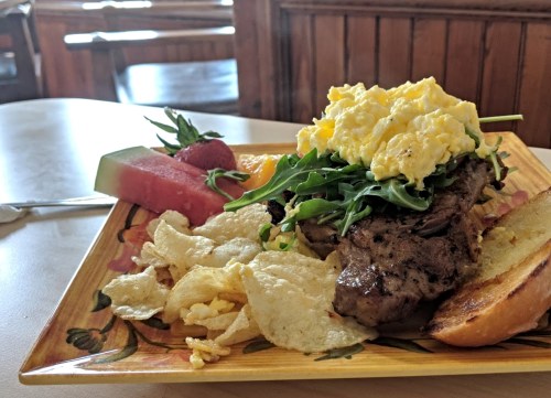 A colorful plate featuring grilled steak topped with scrambled eggs, arugula, chips, and fresh fruit.