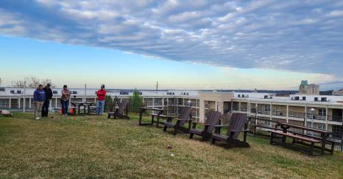 Four people stand on a grassy area with wooden chairs, overlooking a cloudy sky and buildings in the background.