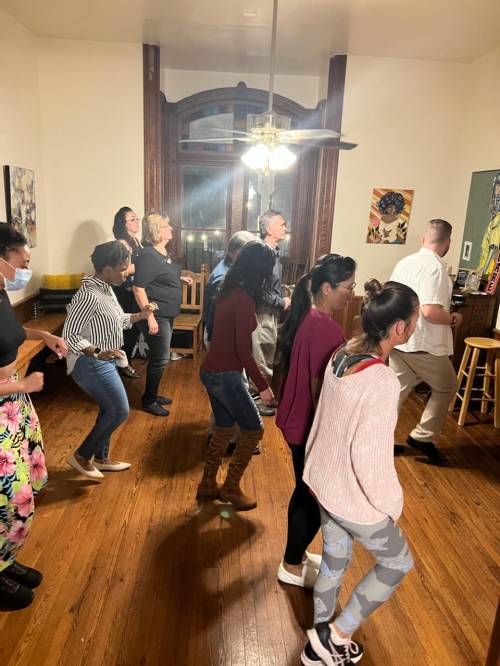 A group of people dancing together in a warmly lit room with wooden floors and a ceiling fan.