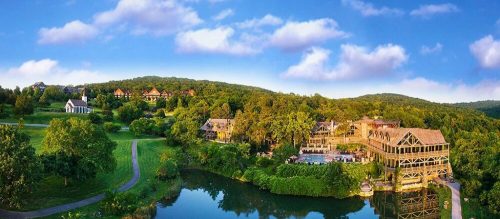 A scenic view of a resort by a lake, surrounded by lush greenery and hills under a blue sky with fluffy clouds.