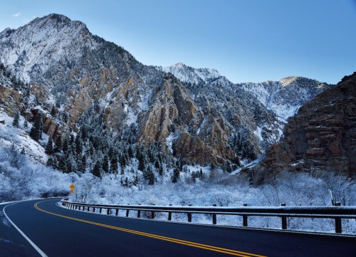 A winding road through snow-covered mountains and trees under a clear blue sky.