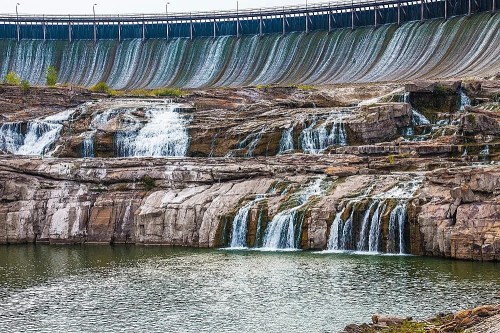 A scenic view of a dam with cascading waterfalls over rocky terrain and a calm body of water below.