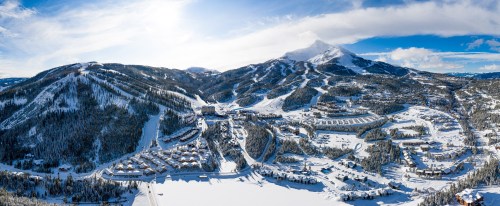 A panoramic view of a snowy mountain landscape with a ski resort and surrounding trees under a clear blue sky.