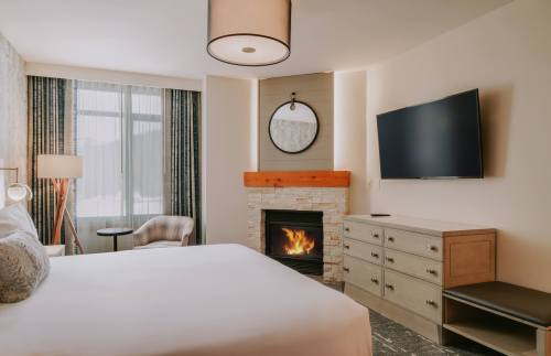 Cozy hotel room featuring a bed, fireplace, TV, and modern decor with large windows and a comfortable chair.
