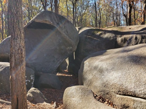 A large boulder rests on smaller rocks in a wooded area, with sunlight creating a rainbow effect on the surface.