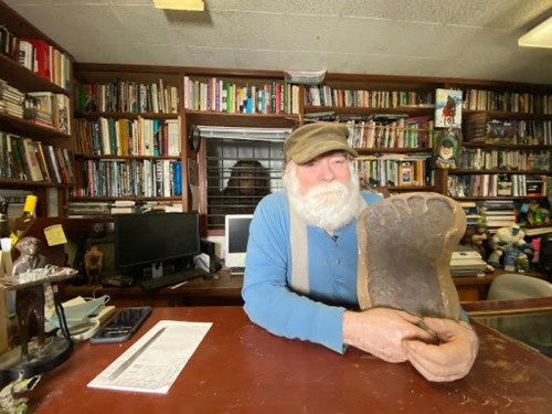 A bearded man in a cap holds a large foot-shaped object, sitting at a desk surrounded by books and a computer.