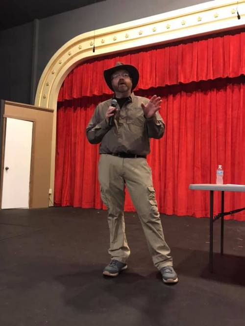 A man in a hat speaks into a microphone on stage, with red curtains in the background and a table beside him.