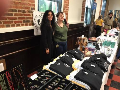 Two women stand behind a table displaying various merchandise, including shirts and jewelry, in a brick-walled venue.