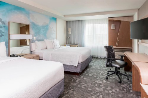 A modern hotel room featuring two beds, a desk, and large windows with light curtains.