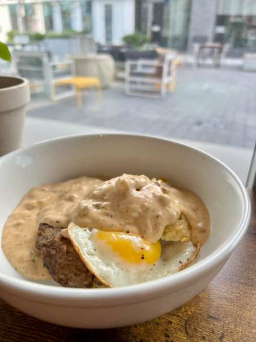 A bowl of creamy sausage gravy over a biscuit topped with a sunny-side-up egg, set against a cafe backdrop.