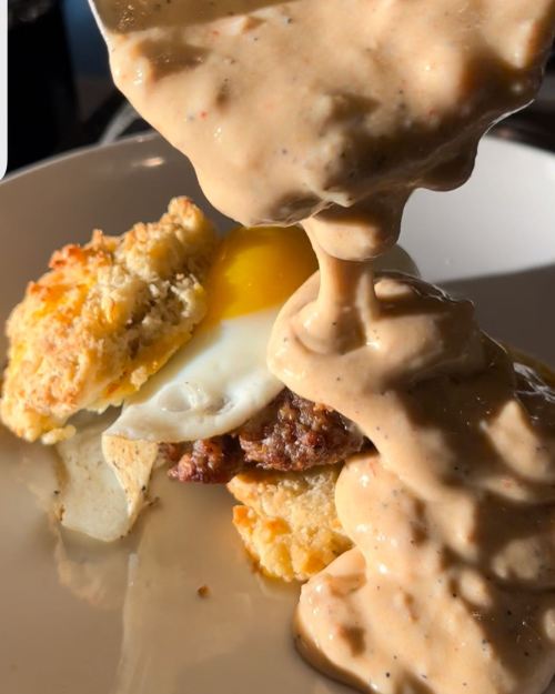 A close-up of a biscuit sandwich with sausage, egg, and creamy gravy being poured over it.