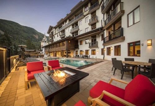 A cozy outdoor area with red chairs, a fire pit, and a pool, surrounded by a mountain view and a multi-story building.