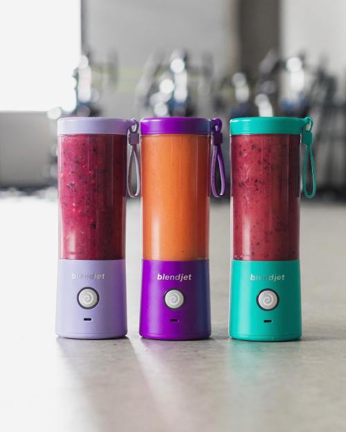 Three BlendJet portable blenders in purple, orange, and teal, filled with colorful smoothies, on a gym floor.