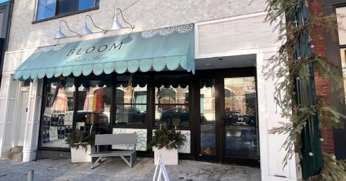 Exterior of a boutique named "BLOOM," featuring a teal awning and decorative planters, on a snowy street.