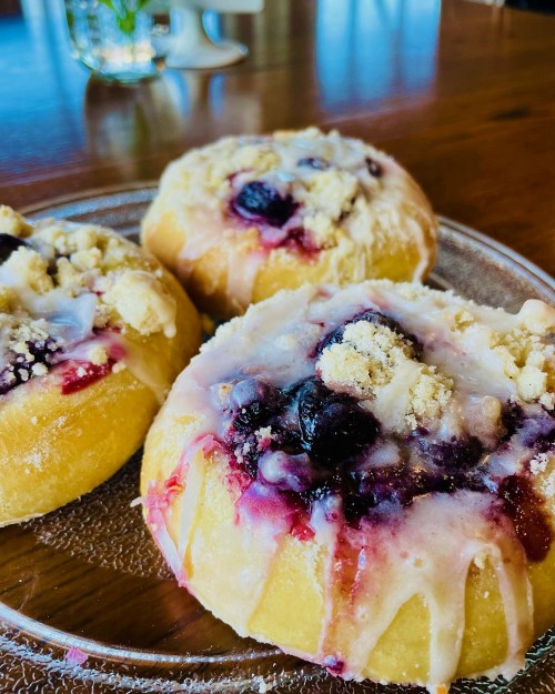 Three sweet pastries topped with blueberries and a crumbly topping, drizzled with icing on a glass plate.