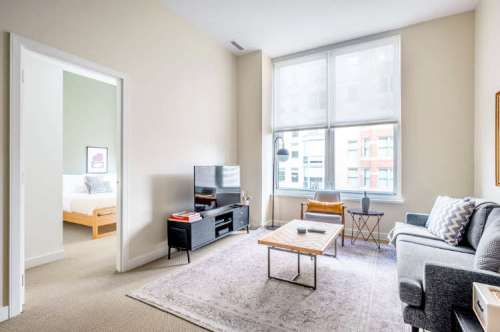 Bright living room with a TV, coffee table, and sofa, featuring large windows and a view of a bedroom through an open door.