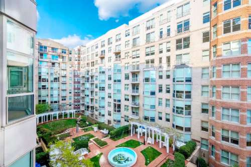 A view of a modern apartment complex courtyard featuring a fountain, greenery, and seating areas under a blue sky.