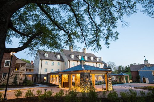 A charming hotel with a stone entrance, surrounded by greenery and trees, illuminated in the evening light.