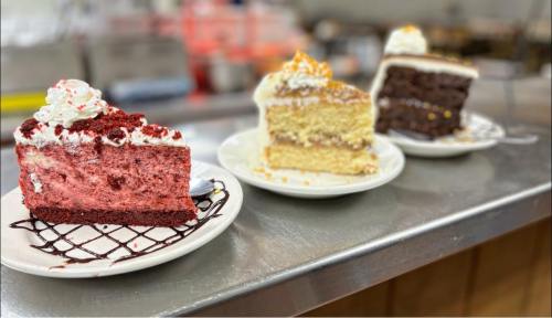 Three slices of cake on plates: red velvet, yellow cake with frosting, and chocolate cake, displayed on a countertop.