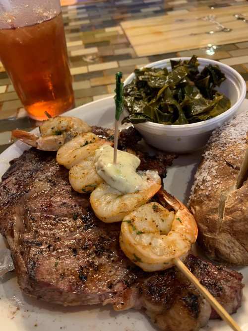 A plate featuring a grilled steak, shrimp skewers with sauce, collard greens, and a baked potato, alongside a drink.