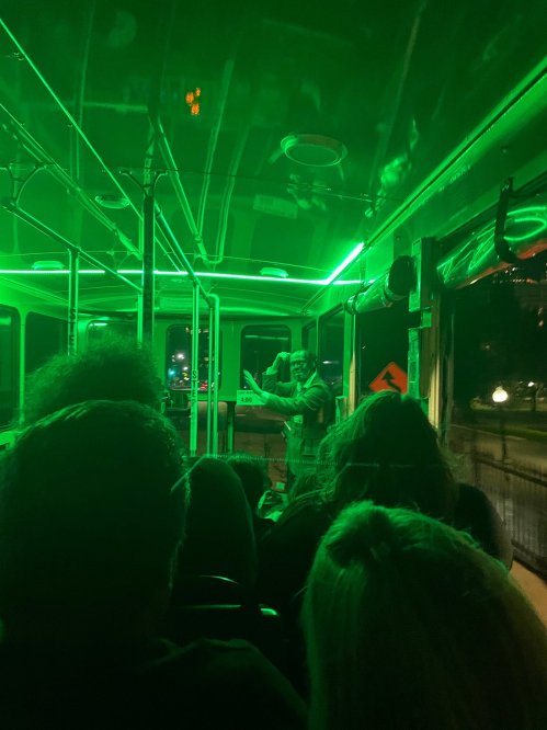 A bus interior illuminated in green light, with passengers seated and a person speaking at the front.