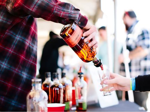 A person pours whiskey from a bottle into a glass at an outdoor event, with other bottles visible in the background.