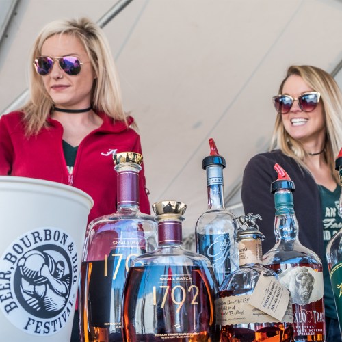 Two women in sunglasses stand behind a table with various whiskey bottles at a beer and bourbon festival.